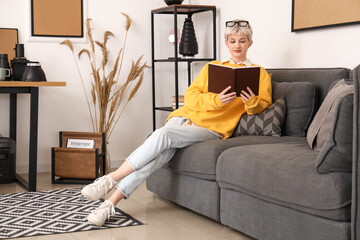 Sticker - Young woman in eyeglasses reading book and sitting on sofa in stylish office