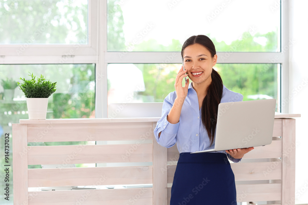 Young female receptionist with modern laptop talking on mobile phone in office