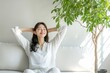 © siripimon2525 - A Japanese woman relaxing on a white sofa, stretching with a smile, indoor plant background. High-resolution, detailed textures, crisp focus