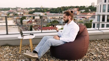 Wall Mural - Man working remotely sitting on rooftop bean bag talking phone with laptop on small table outdoors urban backdrop relaxed work environment