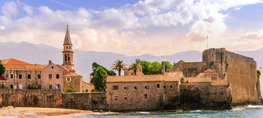 summer season landscape of old town on sea shore with antique wall and tower, blue water vawes and beautiful mountains and cloudy sky on background