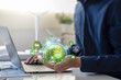 © Antony Weerut - Green Business Sustainable Innovation and Green technology concept. Businessman working on a laptop holds a glowing globe with icons representing renewable energy and sustainable practices.
