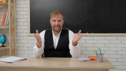 Man college teacher sitting at desk in classroom in front of chalkboard explaining lesson for students. Education concept.