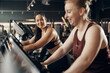 © Flamingo Images - Diverse smiling young woman and a female friend in sportswear riding on stationary bikes during a cardio workout session together at the gym