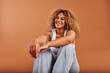 © Flamingo Images - Laughing woman with curly hair, wearing a tank top and jeans sitting on an orange background while she smiles