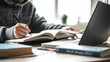 © Anna - Close-up of a person’s hands in a sweater writing in a book at a desk, with a laptop and other books nearby, symbolizing focused study or work.