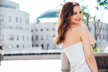 Young beautiful smiling brunette female in trendy evening silver dress. Sexy carefree woman posing in the street at sunny day. Fashionable model with evening bright makeup and red lips, outdoors