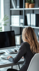 A blonde woman viewed from behind, working at a desk in a modern office with a large monitor, suggesting a tech-savvy and organized professional environment.