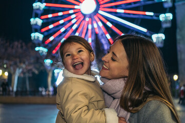 Young Mother’s Candid Moment with Daughter by Illuminated Ferris Wheel. A joyful mother-daughter moment by a lit Ferris wheel.