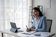 © Wasana - A woman is sitting at a desk with a laptop and a pen