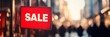 © yanlong - Close-up on a Red Sale Sign on a City Street, with Blurred Shops and People in the Background.