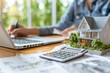 © Yuliia - Beautiful person calculating property tax with a house model on the table.
