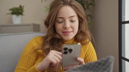 Wall Mural - Young, attractive woman using smartphone indoors, sitting on a couch in a cozy living room.