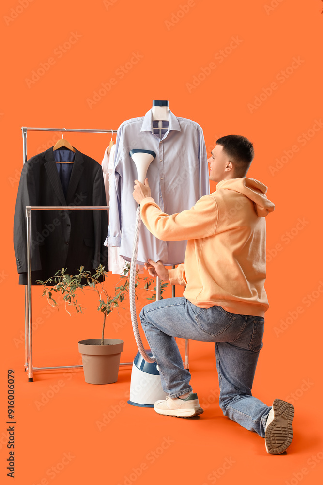 Happy young man steaming clothes on rack on orange background