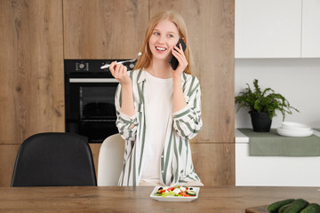 Canvas Print - Young woman talking by mobile phone and eating salad with feta cheese in kitchen