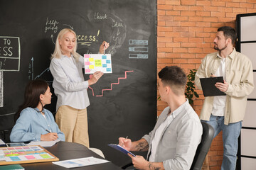 Wall Mural - Mature woman working on business plan near black wall with her colleagues in office