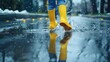 © florynstudio3 - Close-up of yellow rain boots splashing in a puddle of water with rain drops falling around.