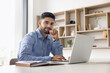 © fizkes - Positive handsome young Arab businessman posing for business portrait at professional workplace, sitting at laptop, typing, touching chin, looking at camera, smiling