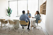 © fizkes - Small business team of three sitting at large meeting table for work discussion, brainstorming on project, planning cooperation. Office corporate white interior, through glass wall view
