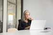 © fizkes - Focused elder company executive woman in suit working at laptop in meeting room alone, sitting at large white office table, typing, using professional application. Business leader candid portrait