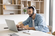 © fizkes - Cheerful handsome Latin freelance entrepreneur man using laptop and headphones for work communication, typing on computer at workplace, smiling, laughing