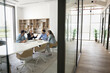 © fizkes - Diverse team of managers meeting for communication in office collaboration room behind glass wall, discussing startup at large table in modern workspace interior. Copy space with hall view