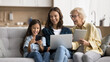 © fizkes - Happy kid, young mother and blonde mature grandmother using gadgets and having fun at home, holding tablet, laptop and smartphone, enjoying Internet technology, communication. Banner shot