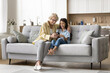 © fizkes - Positive senior grandma and cheerful cute kid girl using online application on mobile phone, sitting on comfortable sofa in cozy home apartment. Granddaughter showing Internet service to granny