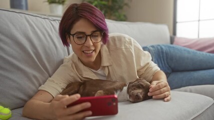 Poster - Young woman with glasses lying on a couch at home with her dog, smiling and using a red smartphone, showcasing a cozy indoor scene.