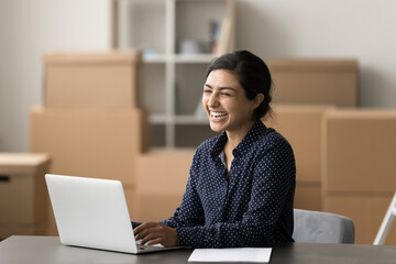 Cheery Indian woman sit at workplace desk laughing working on-line using laptop in warehouse. Small business owner relish first sale or profit growth. Electronic commerce, retailing, merchant workflow