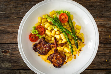 Wall Mural - Lamb chops with bone, gnocchi and cooked green asparagus served on white plate on wooden table
