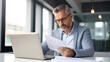 © halo - Portrait of middle aged businessman sitting at desk in office and reading paper document while using laptop.