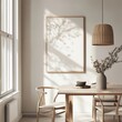 © Bouchra - A white framed picture hangs on a wall in a room with a wooden table and chairs. The table is set with a bowl and a vase of flowers. The room has a simple and clean design
