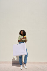 Wall Mural - Happy young African American woman customer wearing sunglasses standing at white wall holding shopping bag using mobile phone making online summer fashion sale purchase in app. Full body vertical shot