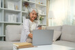 © wattana - Smiling satisfied senior woman with grey hair holding a credit card and using laptop, enjoying online shopping or managing her finances.