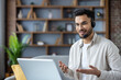 © Tetiana - Indian young man wearing a headset sitting at home in front of a laptop and talking to the camera, gesturing with his hands, smiling at the screen