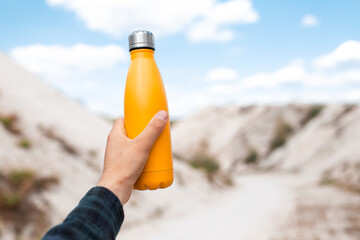 Wall Mural - Male hand holding steel thermo water bottle of yellow color on background of sand hills.