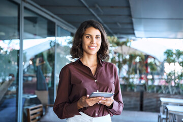 Portrait of young adult professional business woman looking at online trade app. European businesswoman CEO holding digital tablet using fintech tab application, looking at camera in outdoors office