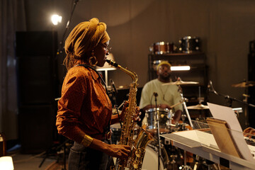 Musicians rehearsing jazz performance with saxophonist in foreground and drummer in background. Recording studio setting with instruments and equipment surrounding musicians