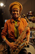 © pressmaster - African American woman standing in recording studio, holding saxophone with bright smile, wearing headwrap and patterned shirt, background showing drum set