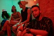 © pressmaster - Young multiethnic man wearing stylish attire vaping and looking at camera while sitting in night club against group of intercultural friends