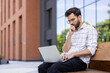 © Tetiana - A young Indian man in casual clothes is sitting on a bench near an office building, holding a laptop on his lap, looking thoughtfully and seriously at the screen