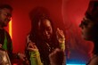 © pressmaster - Young African American woman with long dreadlocks or plaits dancing in front of camera among her friends in neon lit night club