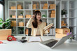 © wichayada - Young Female Entrepreneur Managing Online Business Order in Modern Office with Laptop and Packaging Supplies