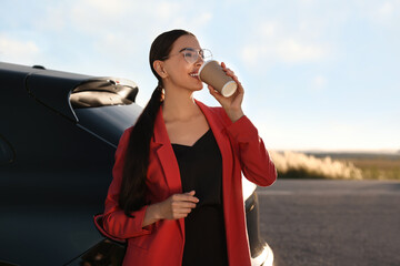 Beautiful young woman drinking from paper cup near car outdoors