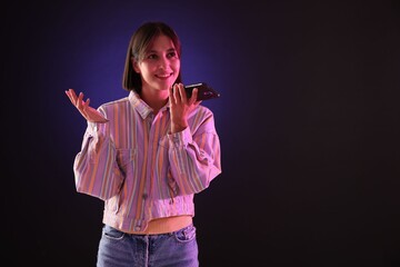 Wall Mural - Beautiful woman recording voice message via smartphone on dark background with neon lights, space for text