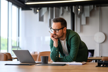 Wall Mural - serious young businessman with hand on chin bending over desk and reading e-mails over laptop in off