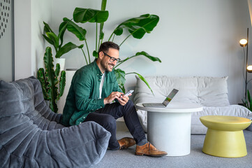 Wall Mural - happy manager with laptop on table using smart phone apps while sitting on sofa in modern workplace