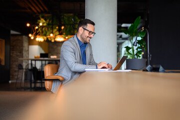 Wall Mural - smiling young businessman in eyeglasses working on his laptop while sitting at desk in modern office