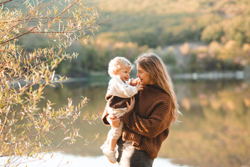 Smiling happy young mother 24-26 year old holding baby girl over lake and nature background. Autumn season. Motherhood.